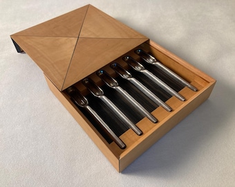 May include: Six stainless steel spoons presented in a wooden box with a geometric lid. The spoons are arranged in a row, and the box is open. The box is made of light-coloured wood. The spoons are for serving.
