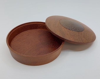 May include: A round wooden box with a lid, both in a warm brown hue. The lid features a circular, textured design. The box is open, revealing the interior. The box is on a white surface.