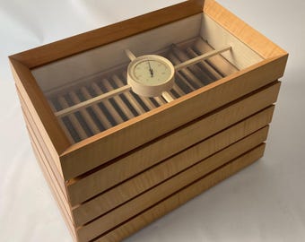 May include: A rectangular wooden humidor with a clear glass top. The humidor is made of light-coloured wood with a layered design. A round hygrometer is visible inside. The humidor is designed to store cigars.