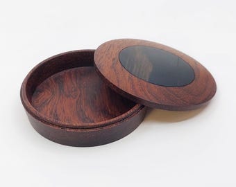 May include: A circular wooden box with a lid, crafted from dark brown wood with a prominent grain. The lid incorporates a black, oval-shaped insert. The box is open, displaying its interior.