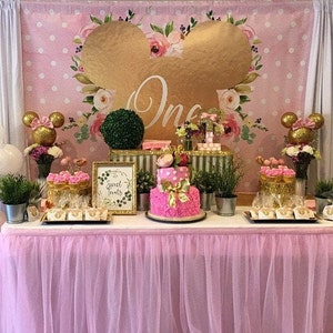 May include: A pink and gold themed first birthday party table setting with a pink and white polka dot backdrop, a gold Mickey Mouse head cutout, a two-tier pink cake with gold accents, and a sign that says "Sweet Treats".