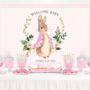 May include: A pink and white gingham backdrop with a watercolor illustration of a bunny wearing a pink jacket. The bunny is surrounded by green leaves and white flowers. The text "Welcome Baby" is above the bunny and the text "Coming June 2021" is below the bunny. The backdrop is set up for a baby shower with pink candy jars, cupcakes, and other decorations.