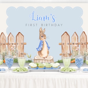 May include: A blue and white backdrop with the text "Liam's First Birthday" and a watercolor illustration of Peter Rabbit standing on a tree stump. The backdrop is decorated with a wooden fence and flowers. There are blue and green candies in glass jars on a white table in front of the backdrop.