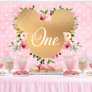 May include: Pink and gold first birthday party backdrop with a gold Mickey Mouse head and floral design. The backdrop has white polka dots and the word "One" is written in gold inside the Mickey Mouse head.