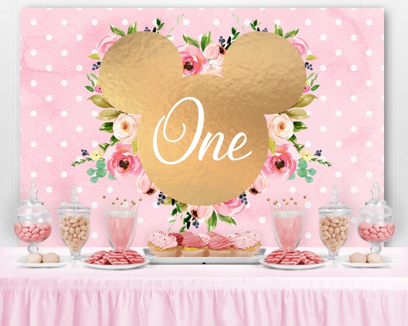 May include: Pink and gold first birthday party backdrop with a gold Mickey Mouse head and floral design. The backdrop has white polka dots and the word "One" is written in gold inside the Mickey Mouse head.