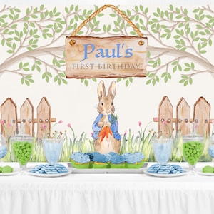 May include: A white backdrop with a green tree branch and a wooden sign that reads "Paul's First Birthday". A watercolor illustration of Peter Rabbit holding a carrot is in the center of the backdrop. A white table with a white tablecloth is in the foreground. There are blue and green candies in glass jars, blue and green cupcakes, and blue and green candies on plates.