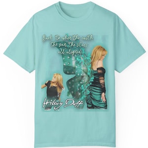 Op de afbeelding: Een lichtblauw t-shirt met een grafische print van een vrouw met een vlinder erachter. De tekst "Back to when the earth, the sun, the stars all aligned..." en "Hilary Duff" staan op het t-shirt gedrukt.