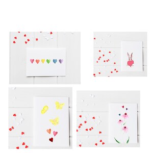 Puede incluir: Cuatro tarjetas de felicitación en blanco con diferentes ilustraciones de acuarela. Las tarjetas están sobre una superficie de madera blanca con confeti de corazones rojos y blancos esparcidos a su alrededor. Las ilustraciones incluyen una fila de corazones arcoíris, una mariposa, un conejito sosteniendo un corazón y una flor rosa.