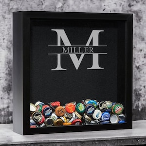 May include: Black shadow box with a collection of beer bottle caps at the bottom. The black background features the name "MILLER" with a large "M" and "IV" in a silver color. The box is displayed on a gray surface.