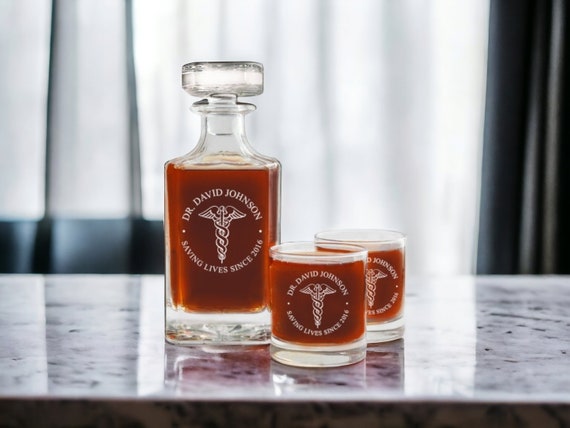 Caduceus-engraved whiskey glasses and decanter set — the medical-symbol personalization is a specific, clever niche that makes a doctor or nurse feel genuinely seen.