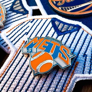 Może przedstawiać: Zbliżenie na przypinkę z koszulki baseballowej New York Mets. Przypinka jest niebieska, biała i pomarańczowa z napisem "Mets" w białym i niebieskim kolorze. Przypinka zawiera również piłkę baseballową w środku.