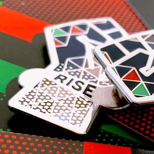 May include: Three silver enamel pins with a raised fist design. The pins are layered on top of each other. The top pin has a black fist with red, green, and white geometric shapes. The middle pin has a black fist with a brown, white, and black geometric pattern. The bottom pin has a white geometric pattern with the word "RISE" in black letters.