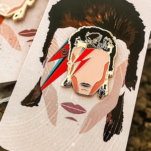 May include: Gold-tone enamel pin featuring a stylized portrait of David Bowie with a red and blue lightning bolt across his face. The pin is on a pink and white background with a swirling pattern.
