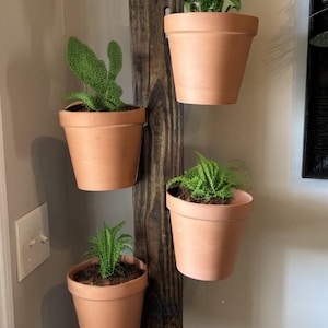 May include: A vertical wooden planter with four terracotta pots, each holding a different green plant. The wood is stained a dark brown, and the pots are suspended from the wood. The planter is hanging on a light-colored wall.
