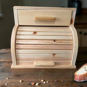 May include: A light-colored wooden bread box with a roll-top door and a drawer. The box is made of natural wood and has a curved design. The drawer and roll-top door have wooden handles. A loaf of bread is visible on the table.