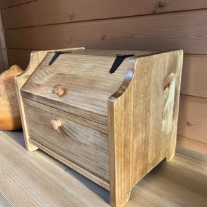 May include: A wooden bread box with a light brown finish. The box has a hinged lid with black metal hinges and a small wooden knob. A drawer with a wooden knob is visible below the lid. The bread box is sitting on a wooden surface.