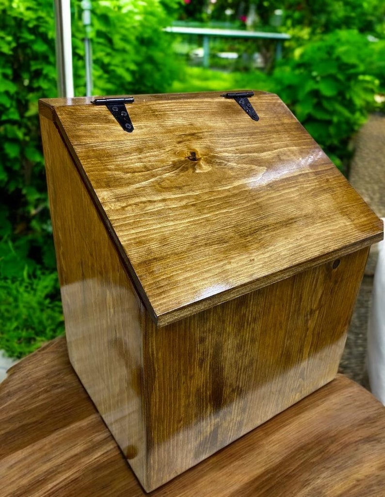 K&ouml;nnte beinhalten: Eine Aufbewahrungsbox aus Holz mit schr&auml;gem Deckel und gl&auml;nzend brauner Oberfl&auml;che, die die nat&uuml;rliche Holzmaserung hervorhebt. Der Deckel ist mit zwei schwarzen Metallscharnieren an der Oberkante befestigt und sorgt f&uuml;r einen sicheren Verschluss.