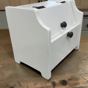 May include: A white wooden storage chest featuring a hinged top lid and a single pull-out drawer. The unit is constructed with smooth white panels and includes two small, round, dark-colored knobs for opening the lid and the drawer.