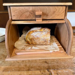 May include: A wooden bread box with a pull-out drawer and a curved design. Inside, a loaf of bread rests on a wooden cutting board and parchment paper. The bread box is made of light brown wood.