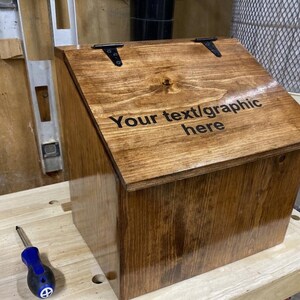 K&ouml;nnte beinhalten: Eine polierte, mittelbraun gebeizte Holzbox mit einem schr&auml;gen, aufklappbaren Deckel, der mit zwei schwarzen Metallscharnieren befestigt ist. Auf dem Deckel ist der schwarze Text "Your text/graphic here" eingraviert. Die Box hat eine glatte, gl&auml;nzende Oberfl&auml;che.
