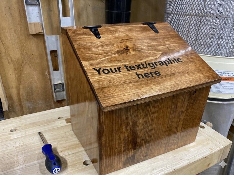 K&ouml;nnte beinhalten: Eine polierte, mittelbraun gebeizte Holzbox mit einem schr&auml;gen, aufklappbaren Deckel, der mit zwei schwarzen Metallscharnieren befestigt ist. Auf dem Deckel ist der schwarze Text "Your text/graphic here" eingraviert. Die Box hat eine glatte, gl&auml;nzende Oberfl&auml;che.