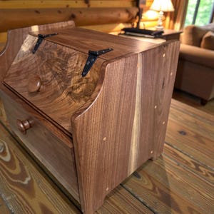May include: A wooden cabinet with a drawer and a hinged top. The cabinet is made of dark brown wood with visible grain patterns and is accented with black hardware. The drawer and top have round wooden knobs.