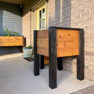 May include: Two wooden raised garden beds with black legs and frames. The beds are made of natural-toned wood and are filled with soil. One bed is in the foreground, and another is in the background. The beds are on a concrete patio.