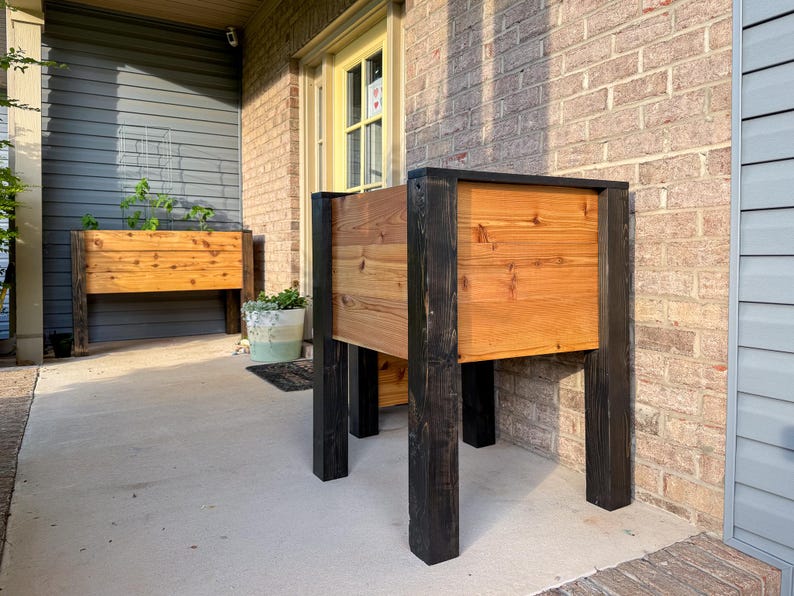 May include: Two wooden raised garden beds with black legs and frames. The beds are made of natural-toned wood and are filled with soil. One bed is in the foreground, and another is in the background. The beds are on a concrete patio.