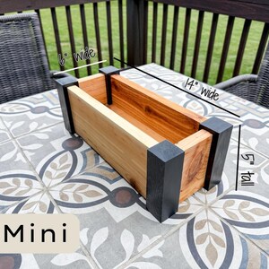 May include: A rectangular wooden planter box, labeled "Mini", with dimensions of 14 inches wide, 6 inches wide, and 5 inches tall. The planter features natural wood tones and black corner accents, suitable for small plants.