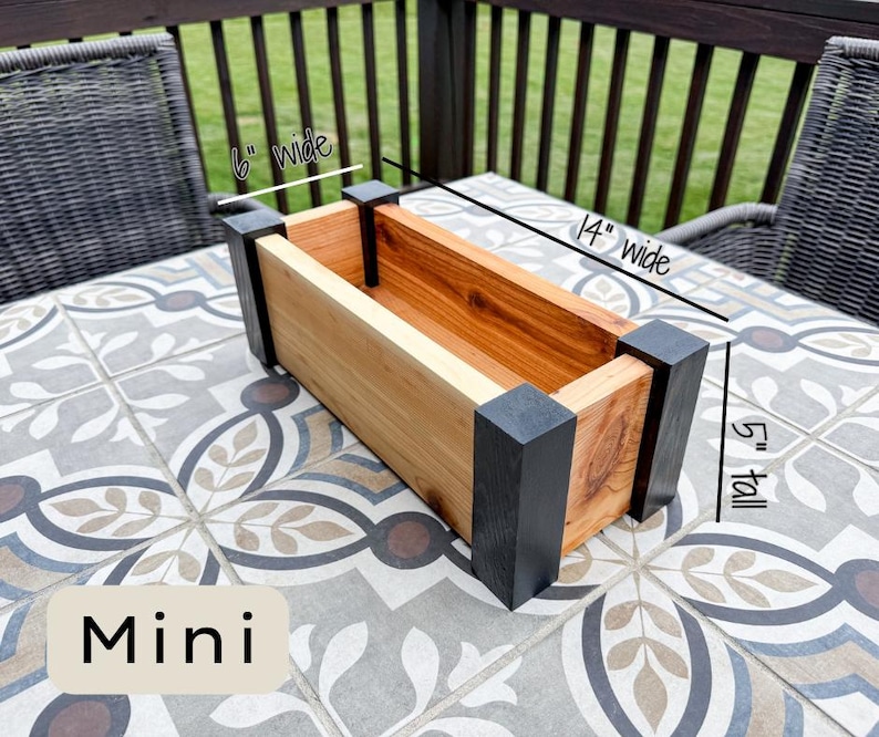 May include: A rectangular wooden planter box, labeled "Mini", with dimensions of 14 inches wide, 6 inches wide, and 5 inches tall. The planter features natural wood tones and black corner accents, suitable for small plants.
