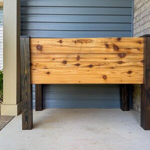 May include: A wooden planter box with a natural wood finish and dark brown legs. The planter box is made of horizontal planks with visible knots. The legs are square and stained a dark brown color. The planter box is designed for outdoor use.