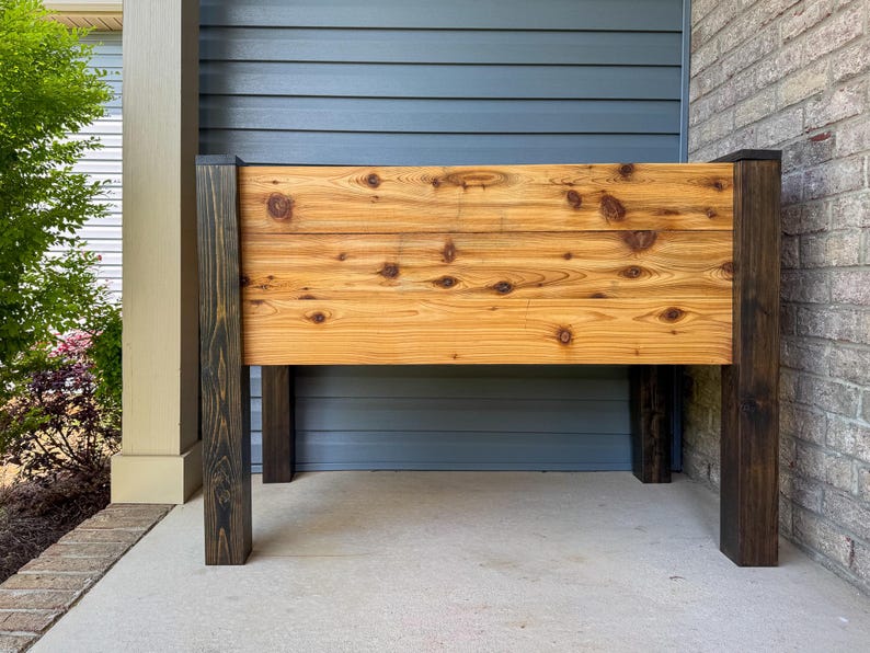 May include: A wooden planter box with a natural wood finish and dark brown legs. The planter box is made of horizontal planks with visible knots. The legs are square and stained a dark brown color. The planter box is designed for outdoor use.