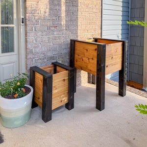 May include: Two wooden raised garden planters with black trim. The planters are made of light-colored wood and have a rectangular shape. One planter is larger than the other. The planters are on a concrete surface.