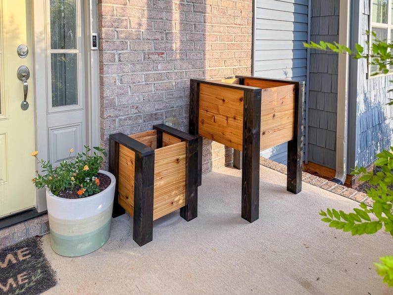 May include: Two wooden raised garden planters with black trim. The planters are made of light-colored wood and have a rectangular shape. One planter is larger than the other. The planters are on a concrete surface.