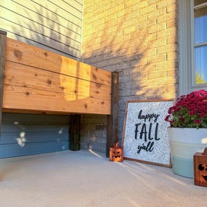 May include: A wooden raised garden bed with dark stained posts. A sign that says "happy FALL y'all" is next to a large pot of red flowers and two carved wooden pumpkins. The scene is set on a porch.