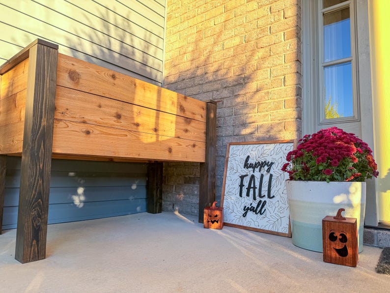 May include: A wooden raised garden bed with dark stained posts. A sign that says "happy FALL y'all" is next to a large pot of red flowers and two carved wooden pumpkins. The scene is set on a porch.
