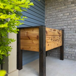 May include: A rectangular wooden planter box with dark stained legs and trim. The planter box is made of light-colored wood with visible knots and grain. The planter is designed for outdoor use and is placed on a concrete surface.
