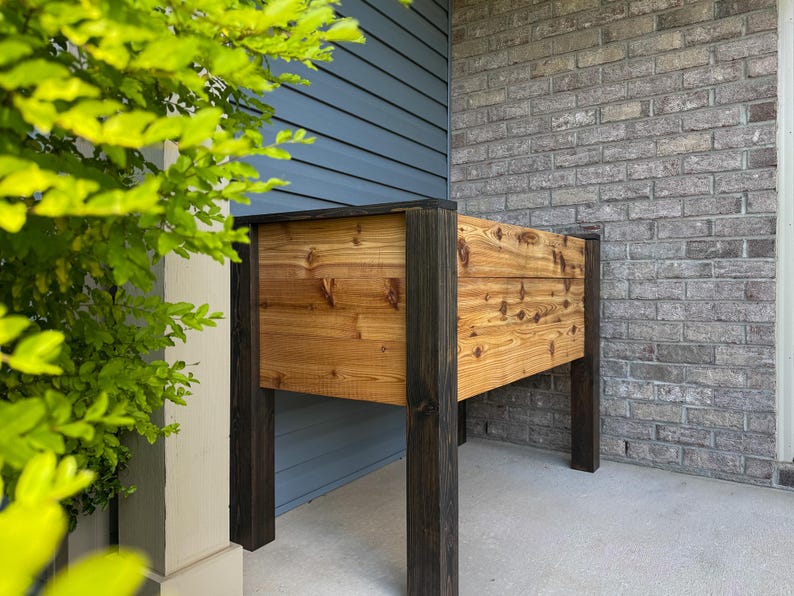 May include: A rectangular wooden planter box with dark stained legs and trim. The planter box is made of light-colored wood with visible knots and grain. The planter is designed for outdoor use and is placed on a concrete surface.