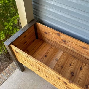 May include: A rectangular wooden planter box with a natural wood finish. The box has dark stained trim and is constructed with visible wood grain and knots. The planter is empty and ready for plants. The background includes a green bush and a gray wall.