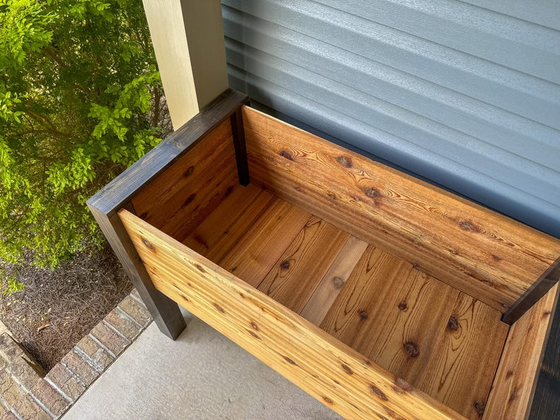 May include: A rectangular wooden planter box with a natural wood finish. The box has dark stained trim and is constructed with visible wood grain and knots. The planter is empty and ready for plants. The background includes a green bush and a gray wall.