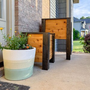 May include: Two wooden planters with a natural wood panel and dark stained frame. A large, round, painted planter with a white, yellow, and green color scheme is next to the planters. The planters are on a concrete porch.