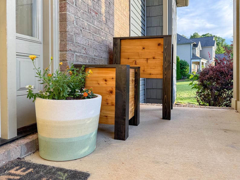May include: Two wooden planters with a natural wood panel and dark stained frame. A large, round, painted planter with a white, yellow, and green color scheme is next to the planters. The planters are on a concrete porch.