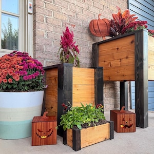 May include: A collection of wooden planters and decorative jack-o'-lanterns. The planters are stained in natural wood tones with black accents. One large planter holds colorful mums. A wooden pumpkin cutout is mounted on the brick wall.