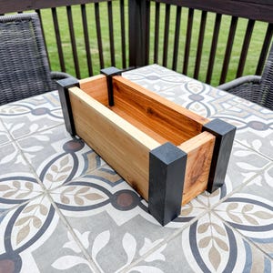 May include: Rectangular wooden planter box with natural and stained wood tones, accented by black corner posts. The planter is empty, showcasing its interior. The box sits on a patterned tile surface.