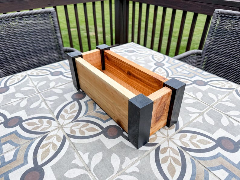 May include: Rectangular wooden planter box with natural and stained wood tones, accented by black corner posts. The planter is empty, showcasing its interior. The box sits on a patterned tile surface.