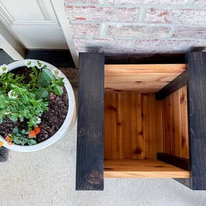 May include: A wooden plant stand with a natural wood interior and dark stained exterior. The stand is next to a white planter filled with green plants and colorful flowers. The stand is made of wood and has a box-like shape.