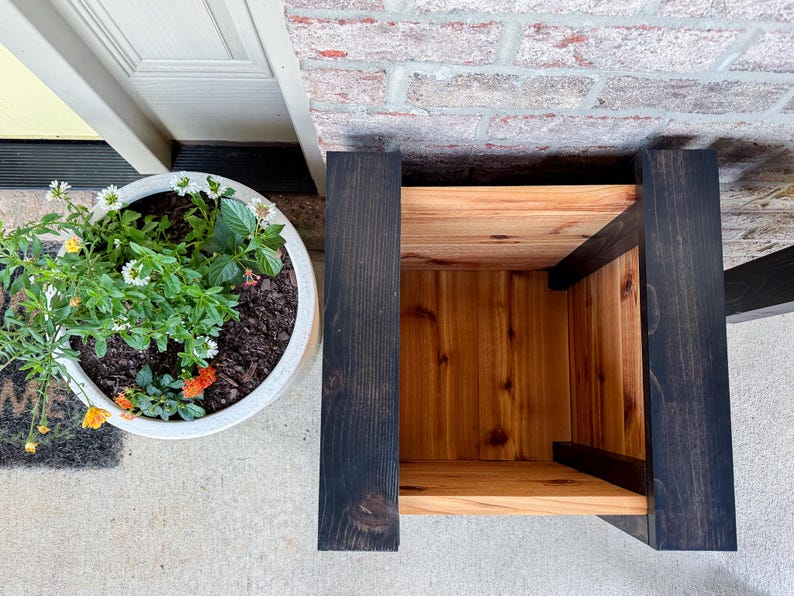 May include: A wooden plant stand with a natural wood interior and dark stained exterior. The stand is next to a white planter filled with green plants and colorful flowers. The stand is made of wood and has a box-like shape.