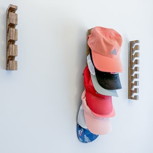 May include: Three vertical wooden wall-mounted racks with multiple notches for storage. One rack holds a stack of six baseball caps in various colors including coral, black, grey, red, light pink, and a blue patterned cap with a white mesh back.