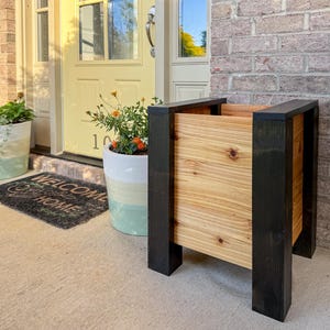 May include: A wooden planter with a natural wood grain center and black painted frame. The planter is rectangular and stands on four legs. The background includes a front door, welcome mat, and potted plants.