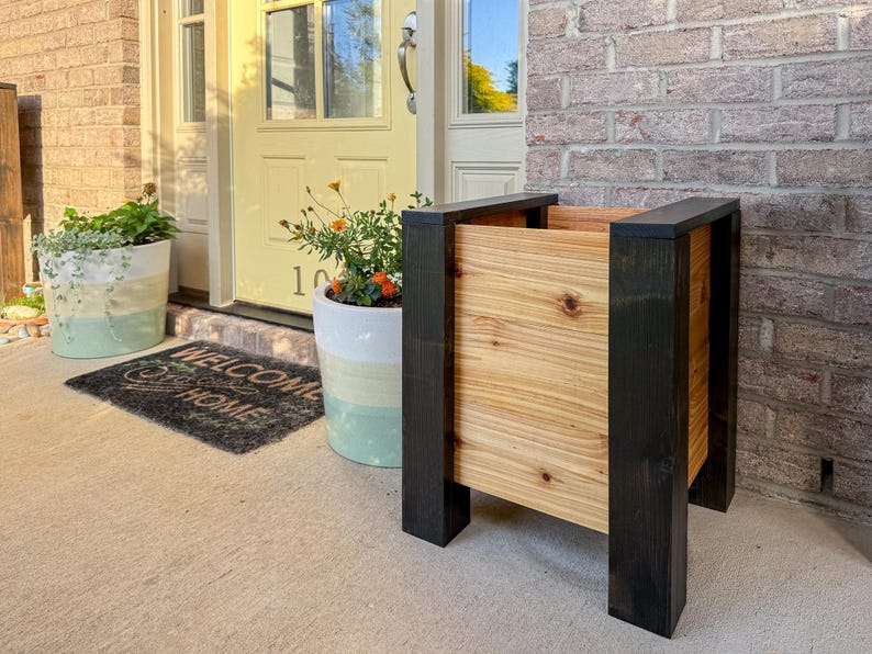 May include: A wooden planter with a natural wood grain center and black painted frame. The planter is rectangular and stands on four legs. The background includes a front door, welcome mat, and potted plants.